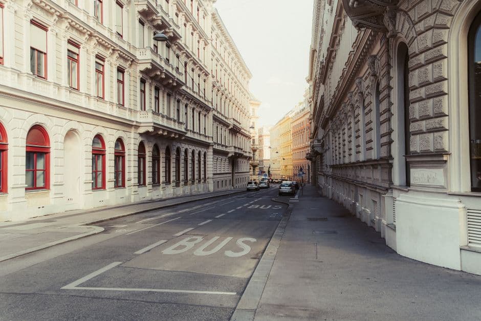 A quiet Vienna side street lined with pale Historicist apartment buildings with red-framed windows, a bus lane marked on the road and parked cars in the distance