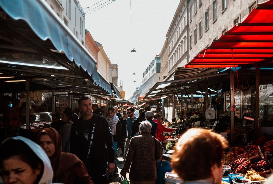 A crowded market lane in Vienna lined with permanent awning-covered stalls selling fruit and vegetables, with pale historic apartment buildings rising at the end of the street