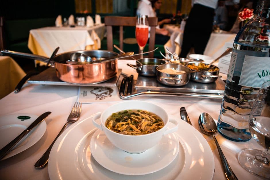 A restaurant table set with a copper pot of beef broth on a warming stand, small copper and steel pans of sauces beside it, and a white two-handled cup of clear broth with sliced pancake strips in the foreground