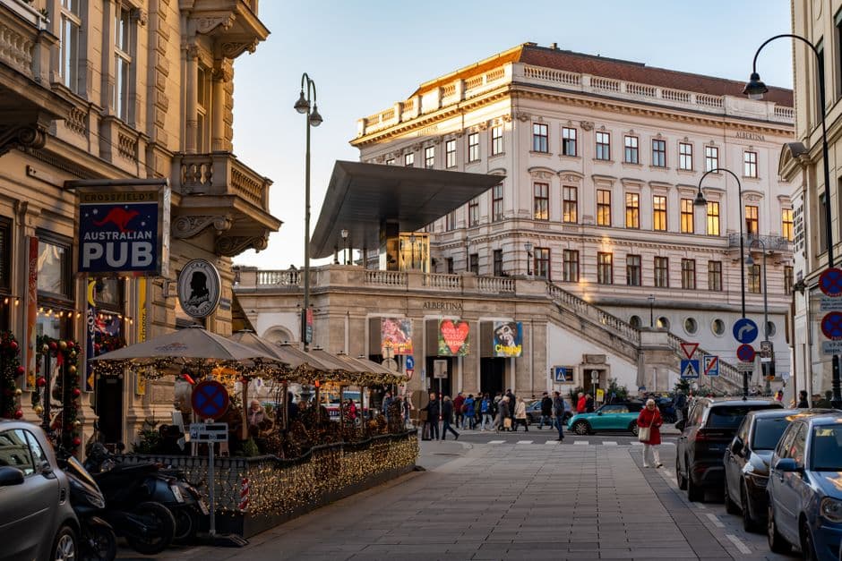 A street in central Vienna in front of the Albertina museum, with a cafe terrace strung with lights in the foreground and people crossing the road