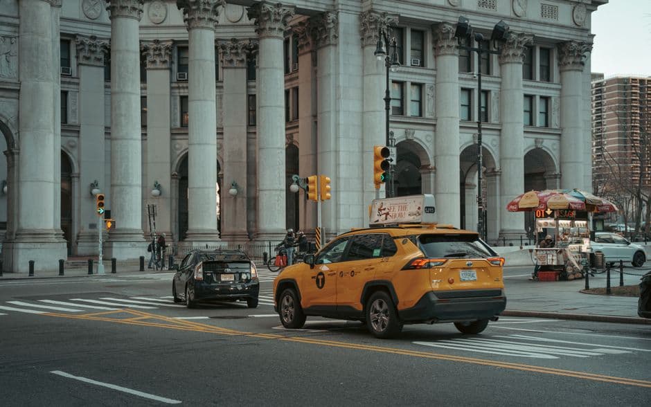 A yellow New York City taxi crossing an intersection in front of a columned stone building, with a street food cart under a striped umbrella on the corner