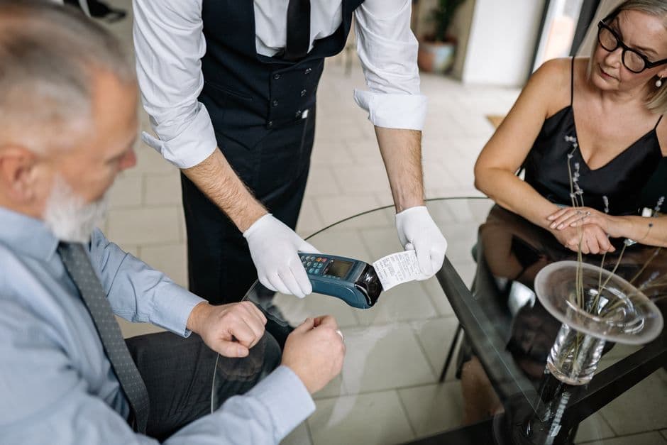 A waiter holding a card payment terminal and a printed receipt out to a couple at a restaurant table