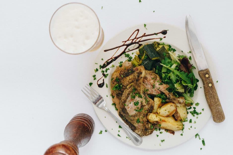 A grilled pork chop with roast potatoes and salad leaves on a white plate, next to a glass of pale lager