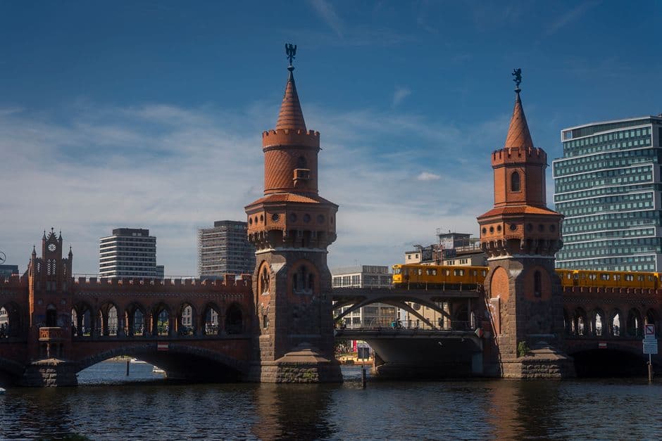 The Oberbaum Bridge over the Spree in Berlin, with its two red-brick towers and a yellow U-Bahn train crossing the upper deck under a blue sky