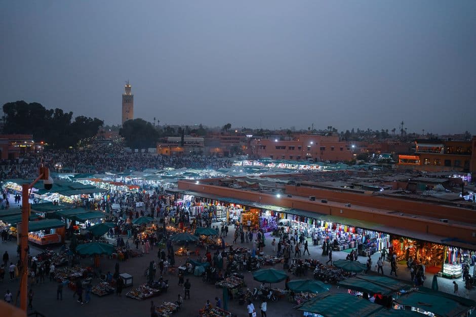 Jemaa el-Fnaa square in Marrakech at dusk, seen from above, with rows of lit market stalls under green awnings, crowds, and the Koutoubia minaret on the skyline